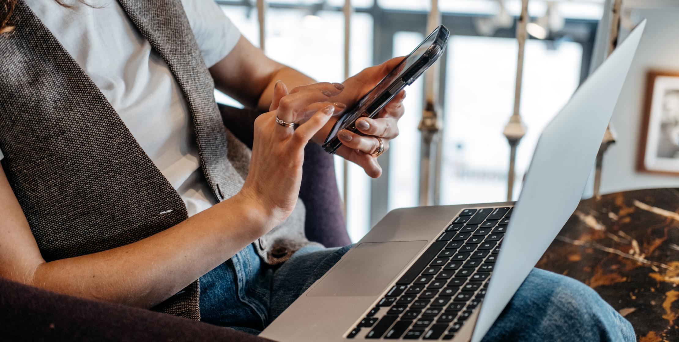 Person sitzt mit Laptop auf dem Schoß und tippt etwas auf ihrem Handy ein