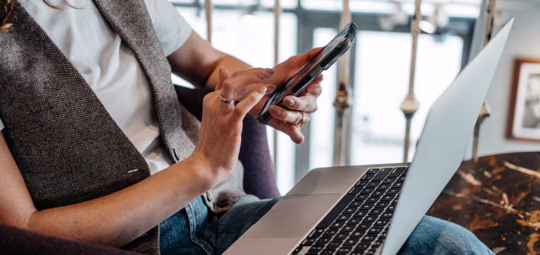 Person sitzt mit Laptop auf dem Schoß und tippt etwas auf ihrem Handy ein